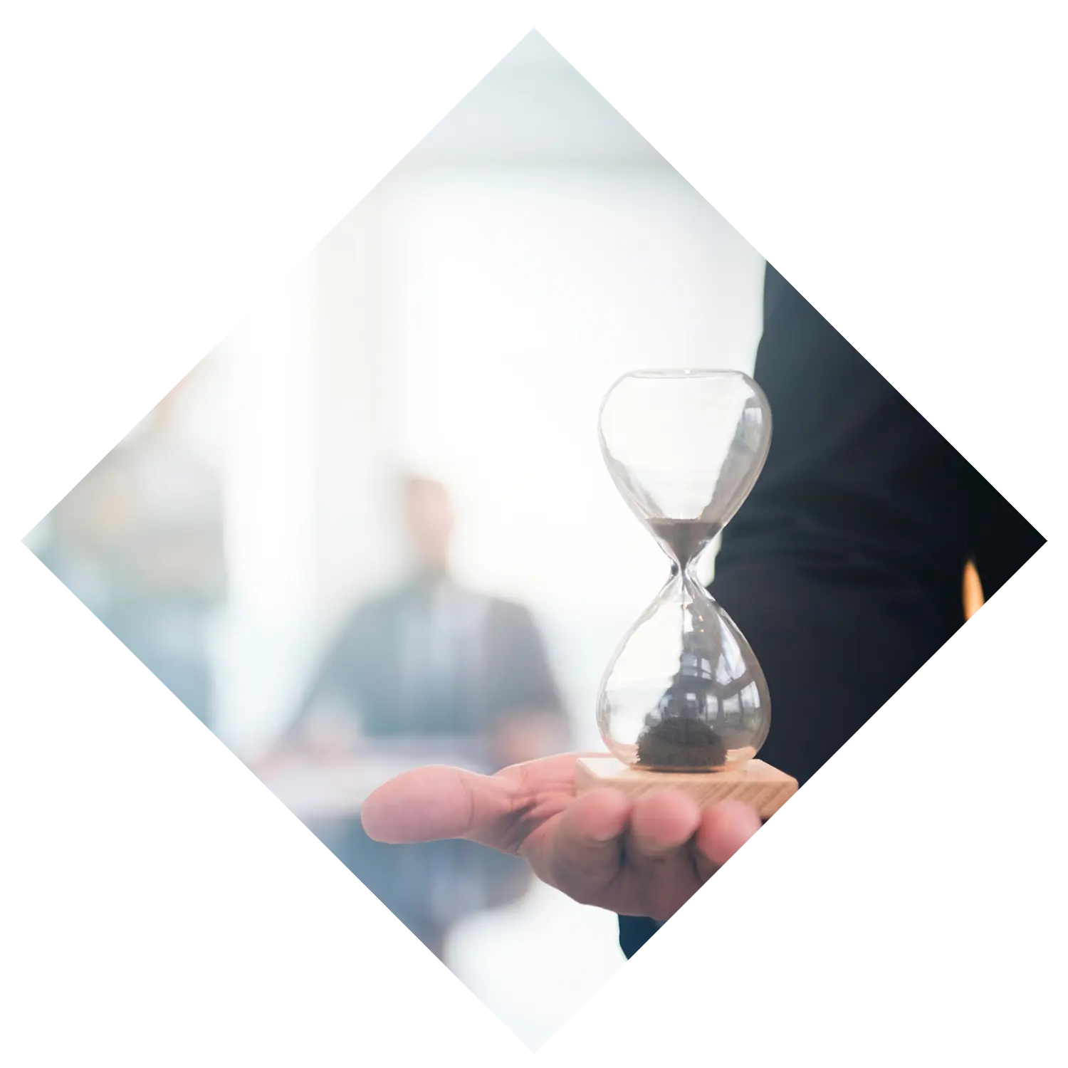 Image of a businessman's hand holding an hour glass with other business people seated at a table behind him representing the duration of time you require a consultant's services.