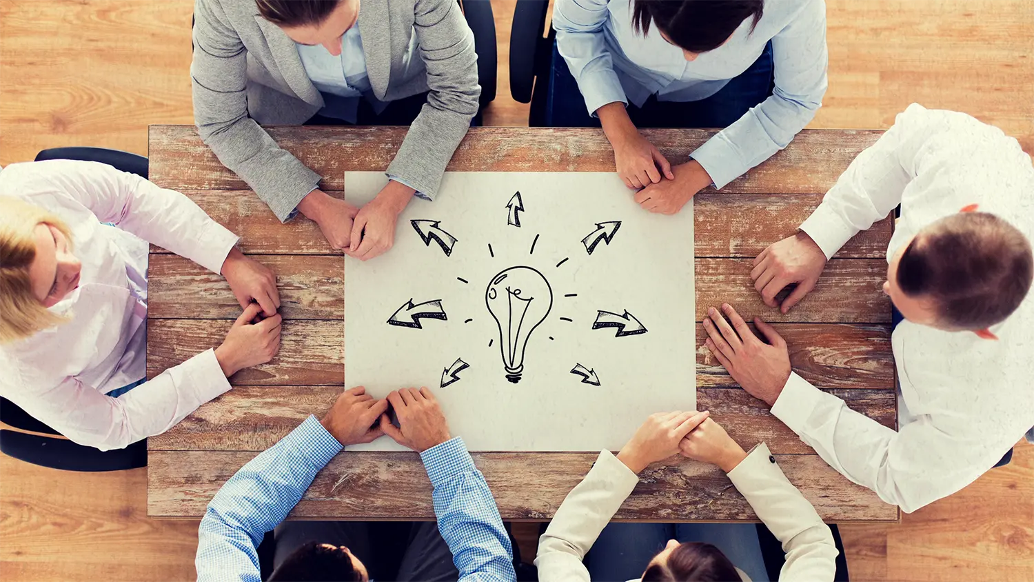 Image of 6 business people sitting around a table with a paper in the center featuring a lightbulb sketch with arrows  pointing in different directions representing talking to a business consultant.